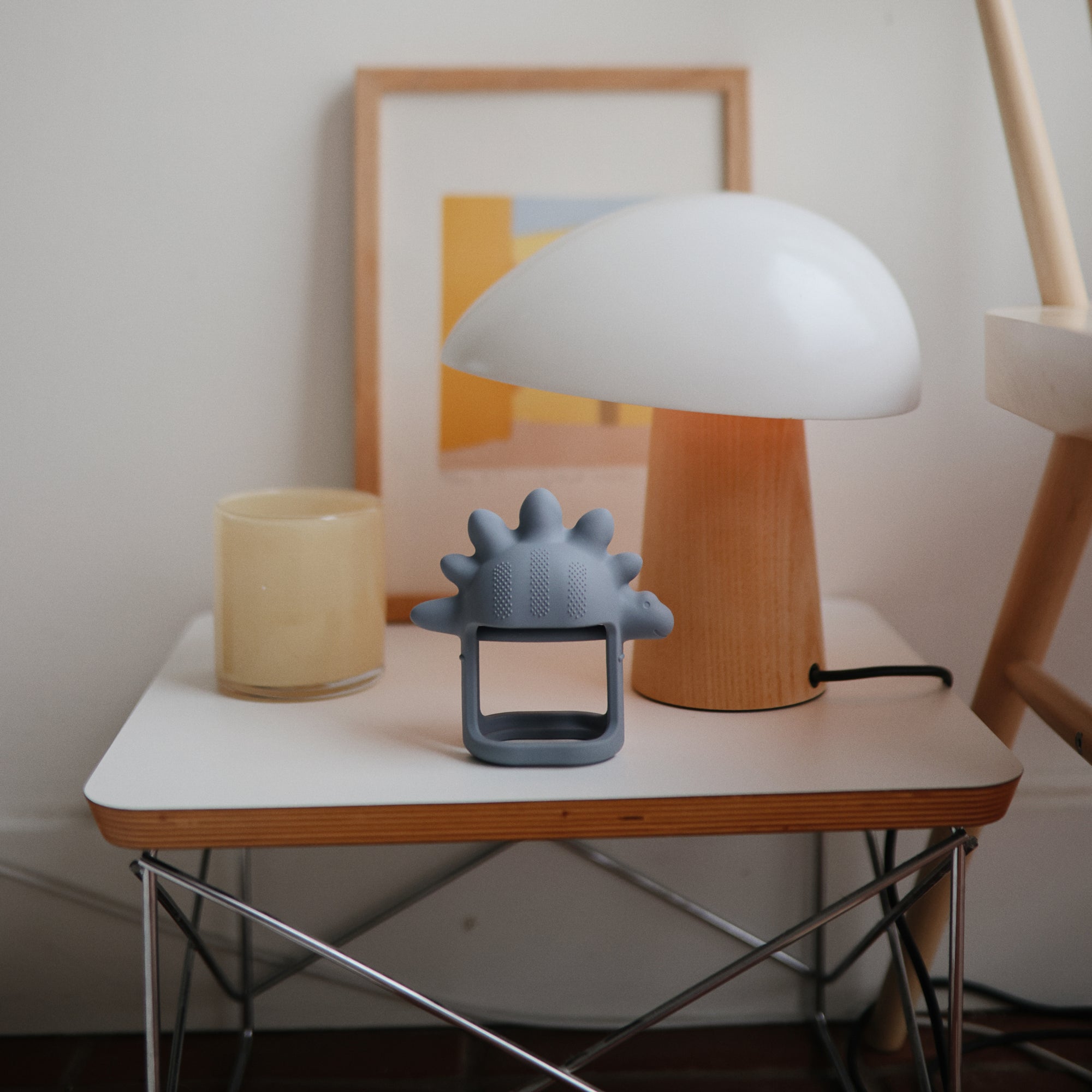 A modern lamp, framed art, and a mushie No-Drop Teether sit on a small white table.