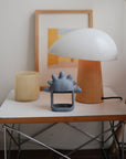 A modern lamp, framed art, and a mushie No-Drop Teether sit on a small white table.