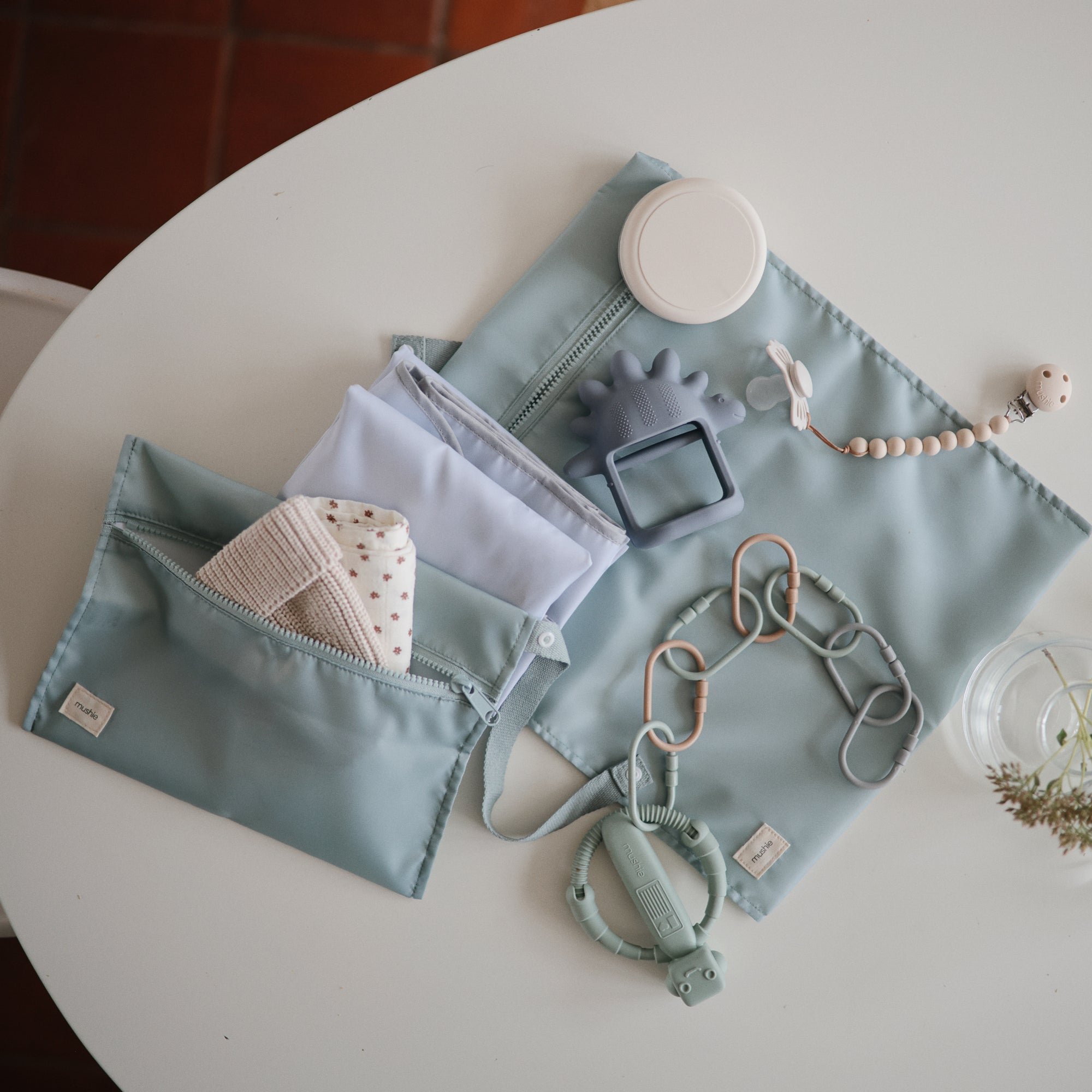 Mushie No-Drop Teether, pacifier clip, and soft cloths in blue pouches on a table.