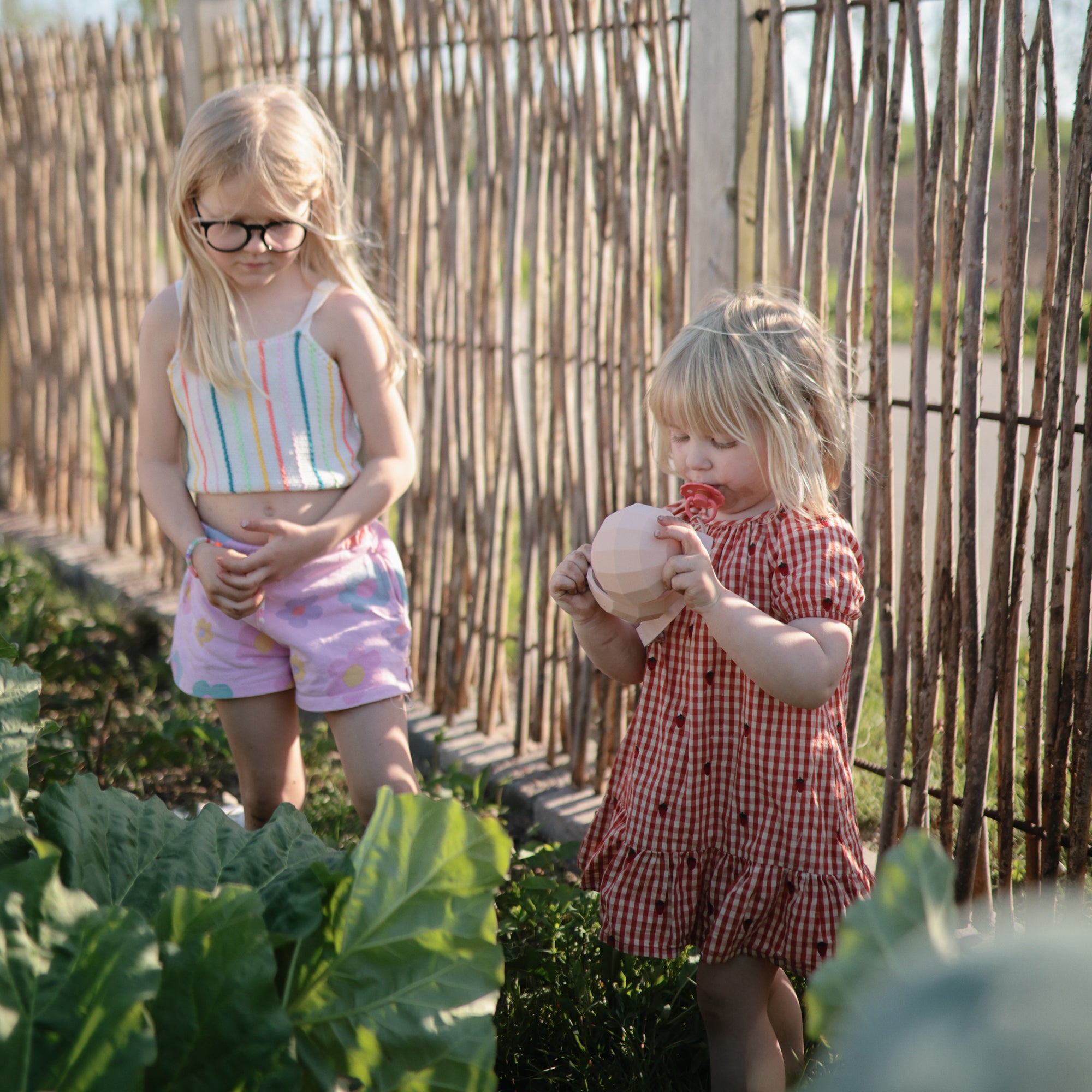 Two girls in a garden; one enjoys fruit holding a mushie Nesting Spheres Sensory Toy.