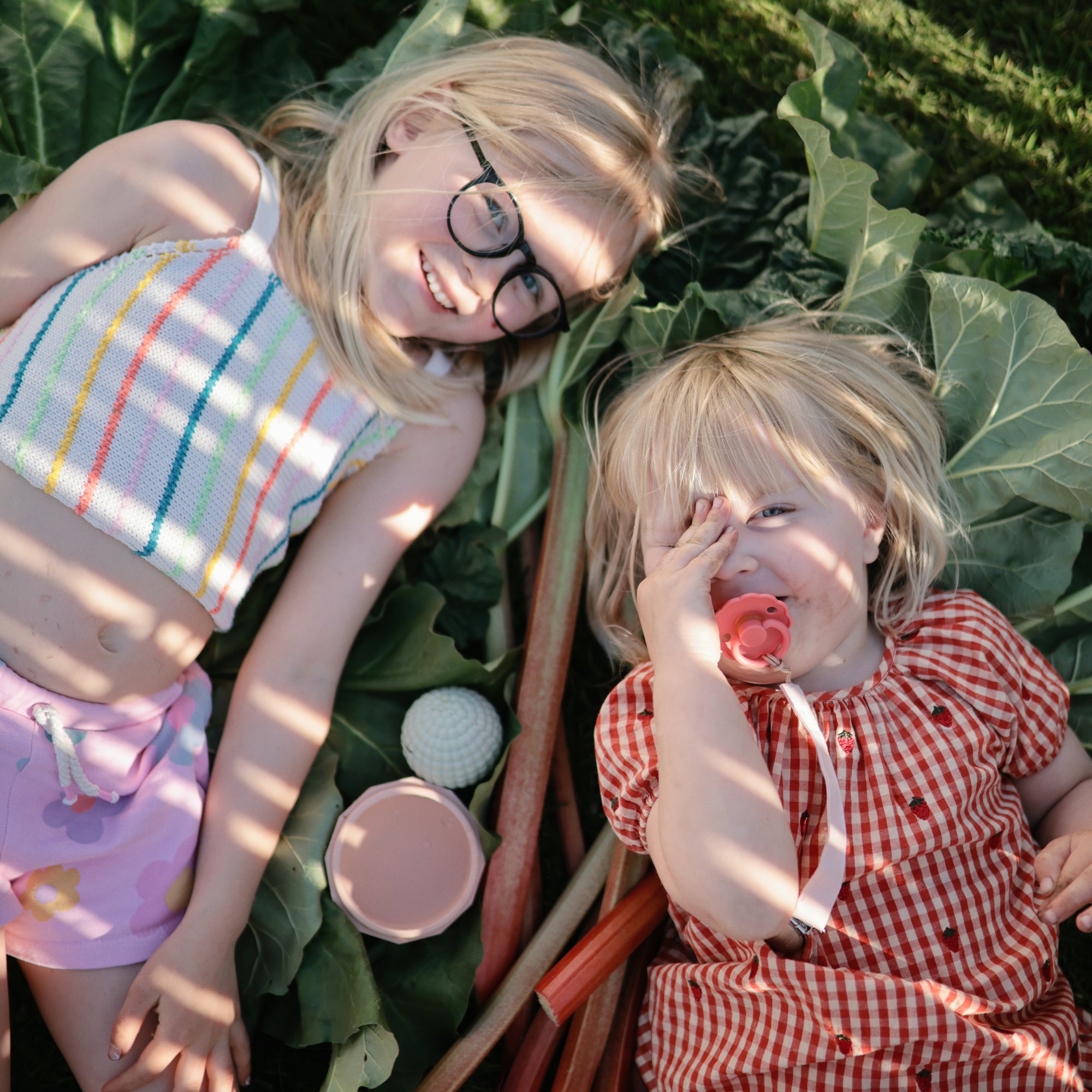 Two kids enjoy the mushie Nesting Spheres Sensory Toy, smiling while sitting on green leaves.