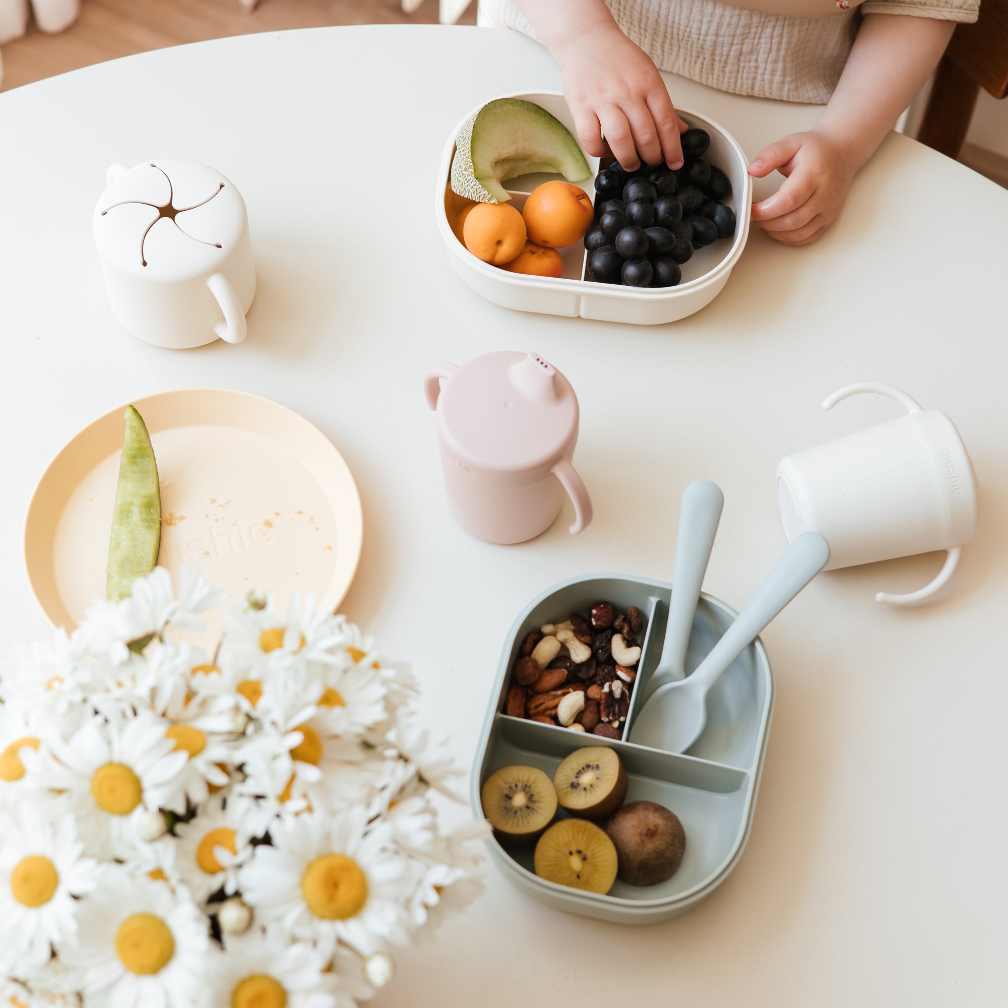 Child’s hands with snacks, plates, and a mushie Dinnerware Fork and Spoon Set on the table.