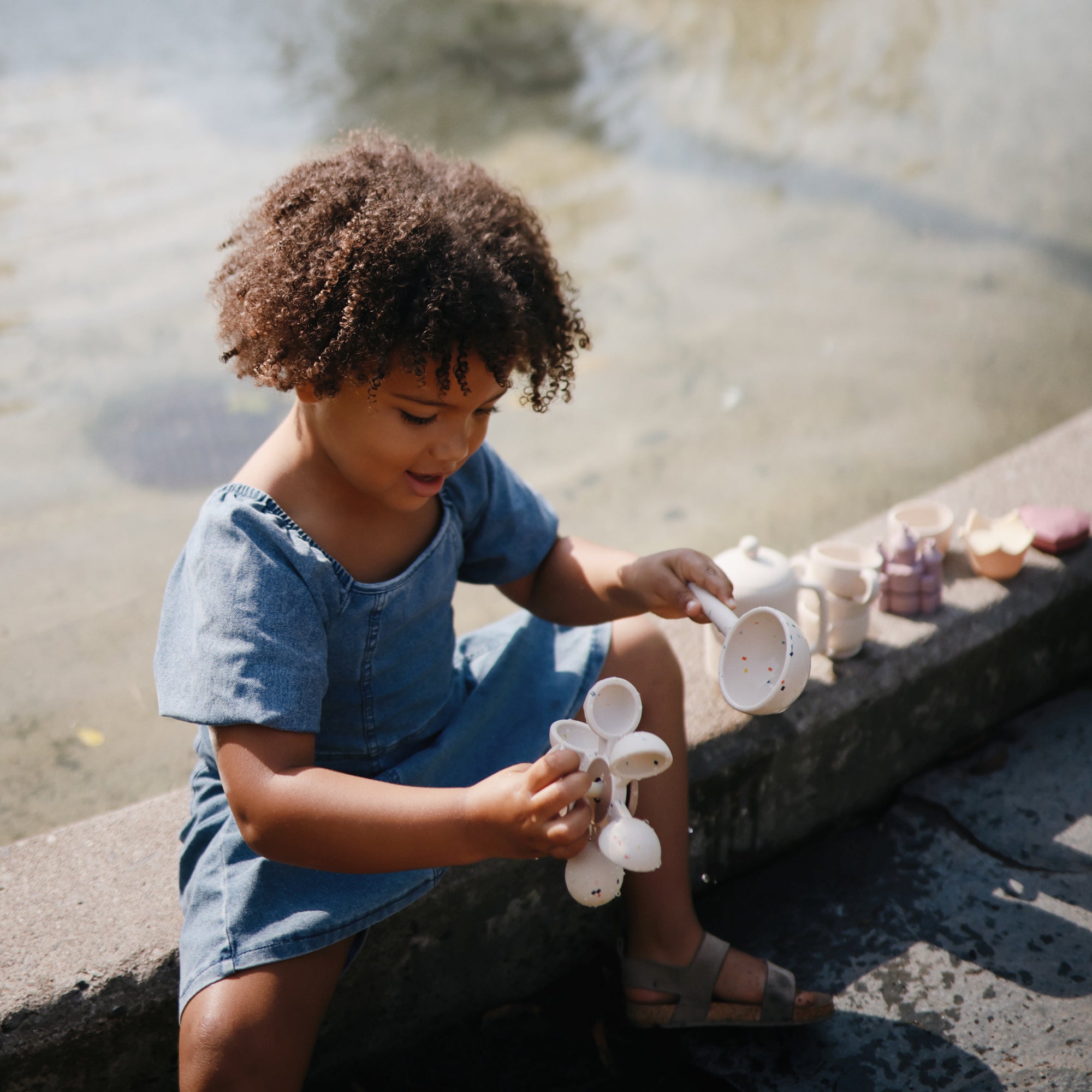Young child plays outdoors by water with mushie's Water Wheel Bath Toy in blue.