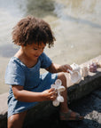 Young child plays outdoors by water with mushie's Water Wheel Bath Toy in blue.