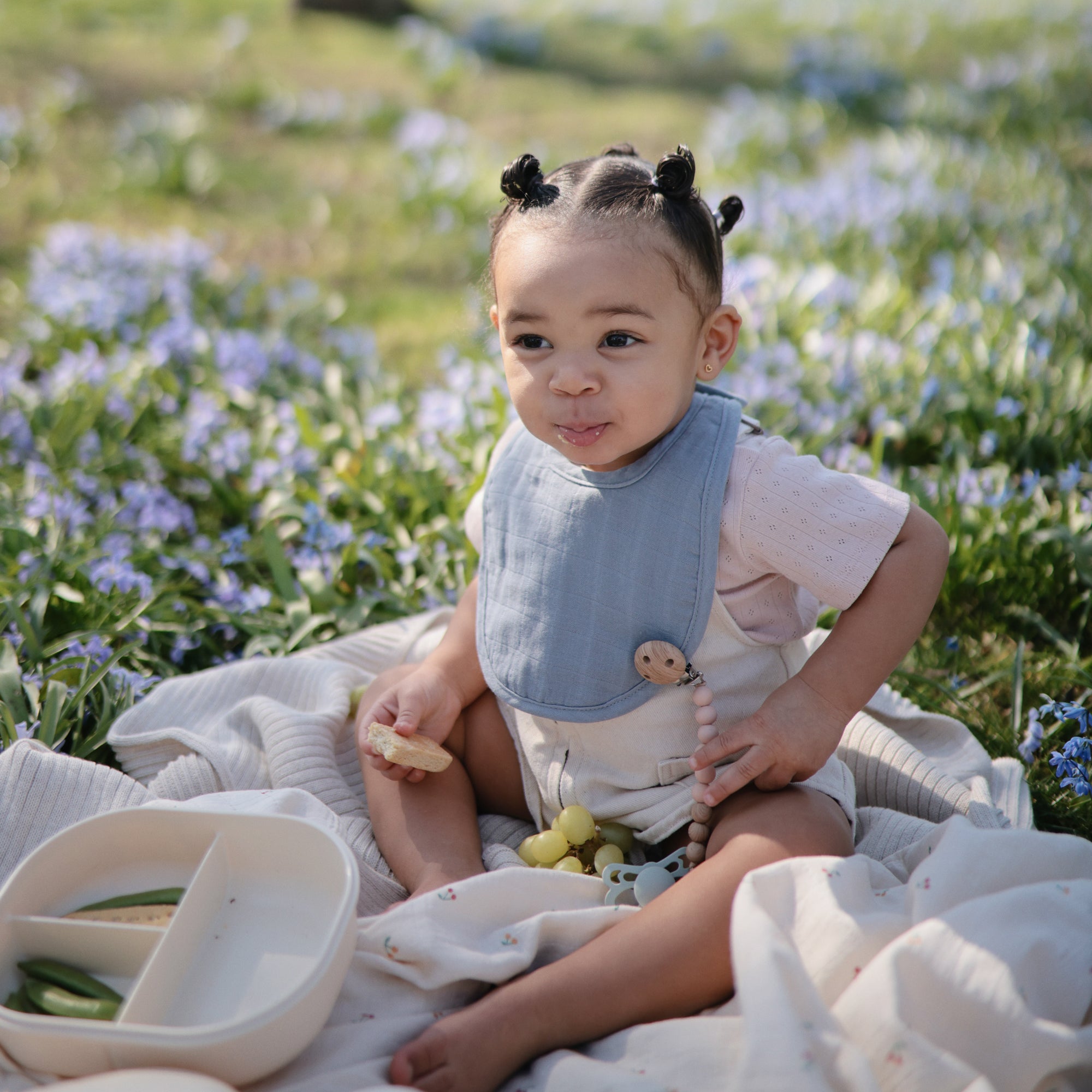 Toddler enjoys a snack from a mushie Lunchbox with compartments, amid grass and flowers.