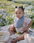 Toddler enjoys a snack from a mushie Lunchbox with compartments, amid grass and flowers.