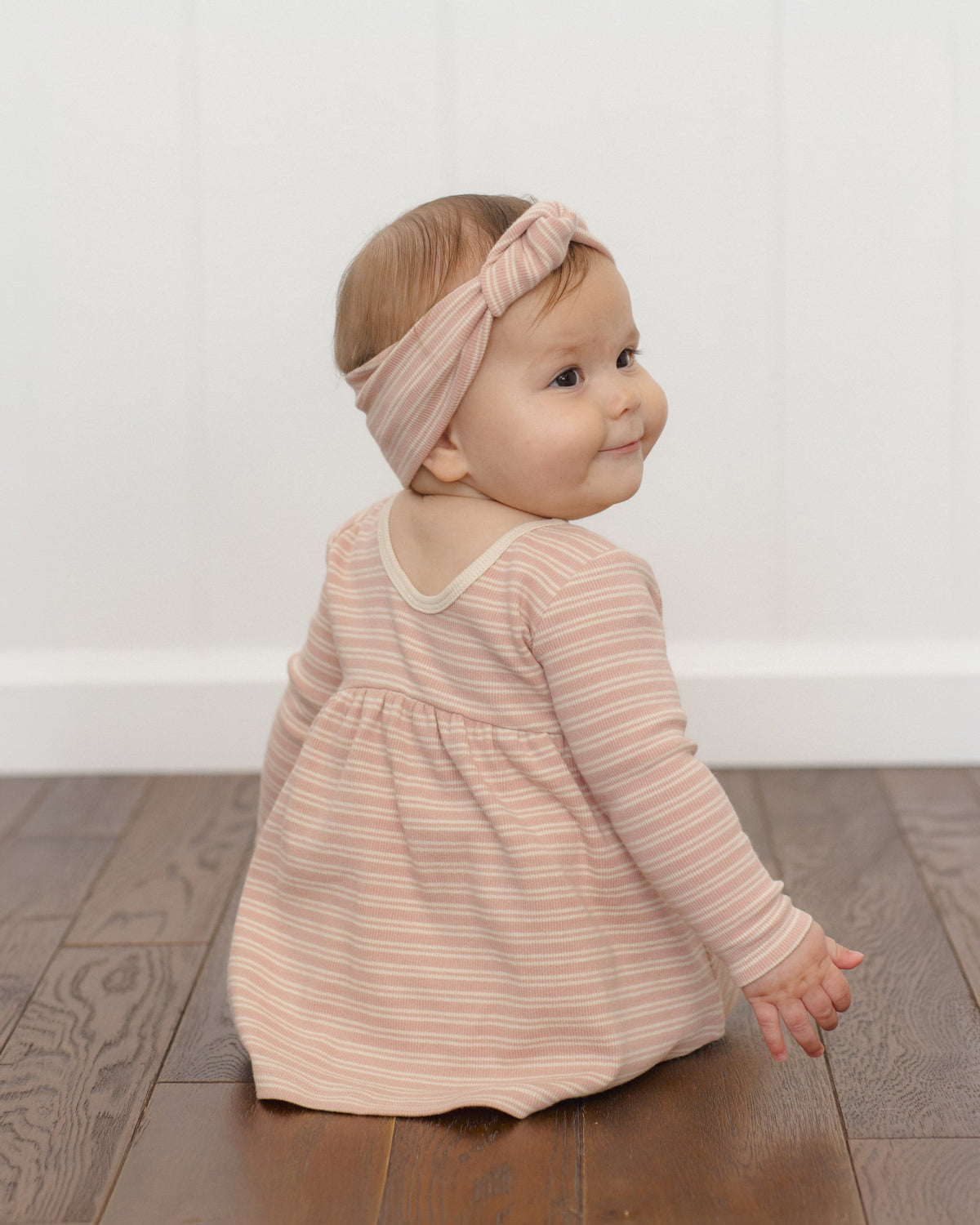 Baby in a Rylee + Cru Ribbed Long Sleeve Dress in Dusty Rose Microstripe sits, looking back.
