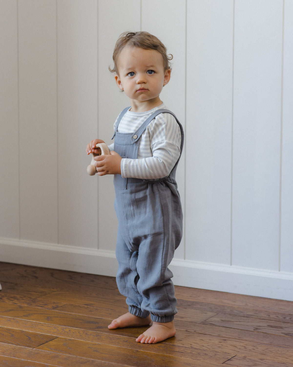 Toddler in gray overalls and Rylee + Cru Long Sleeve Tee Cloud Stripe stands barefoot with toy.