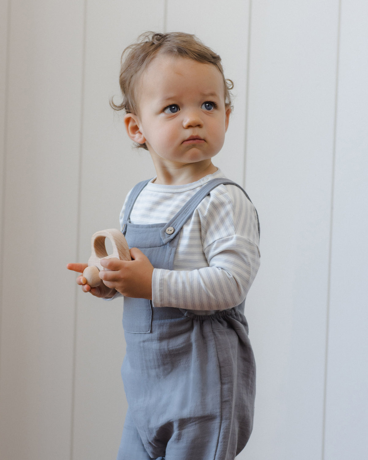 Toddler in blue overalls and Rylee + Cru Long Sleeve Tee Cloud Stripe by a white paneled wall.