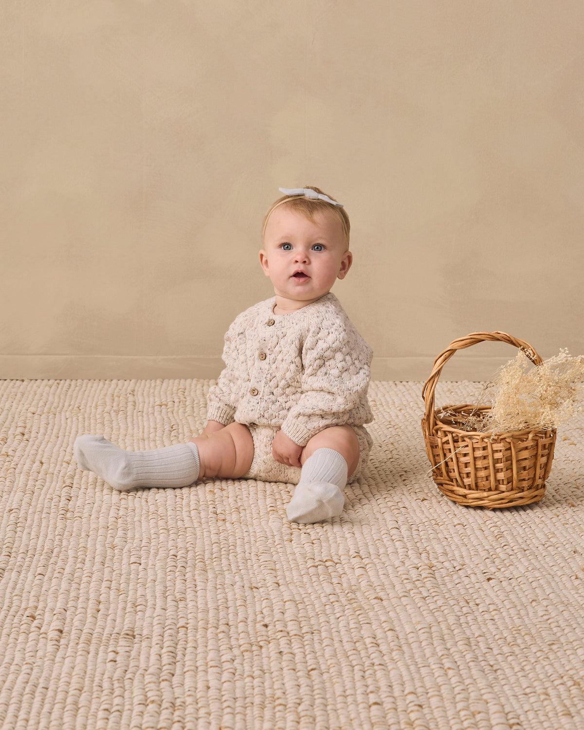 Baby in Rylee + Cru Knit Bloomer Natural Confetti sits on woven rug by a wicker basket.