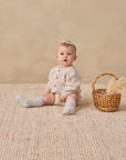 Baby in Rylee + Cru Knit Bloomer Natural Confetti sits on woven rug by a wicker basket.