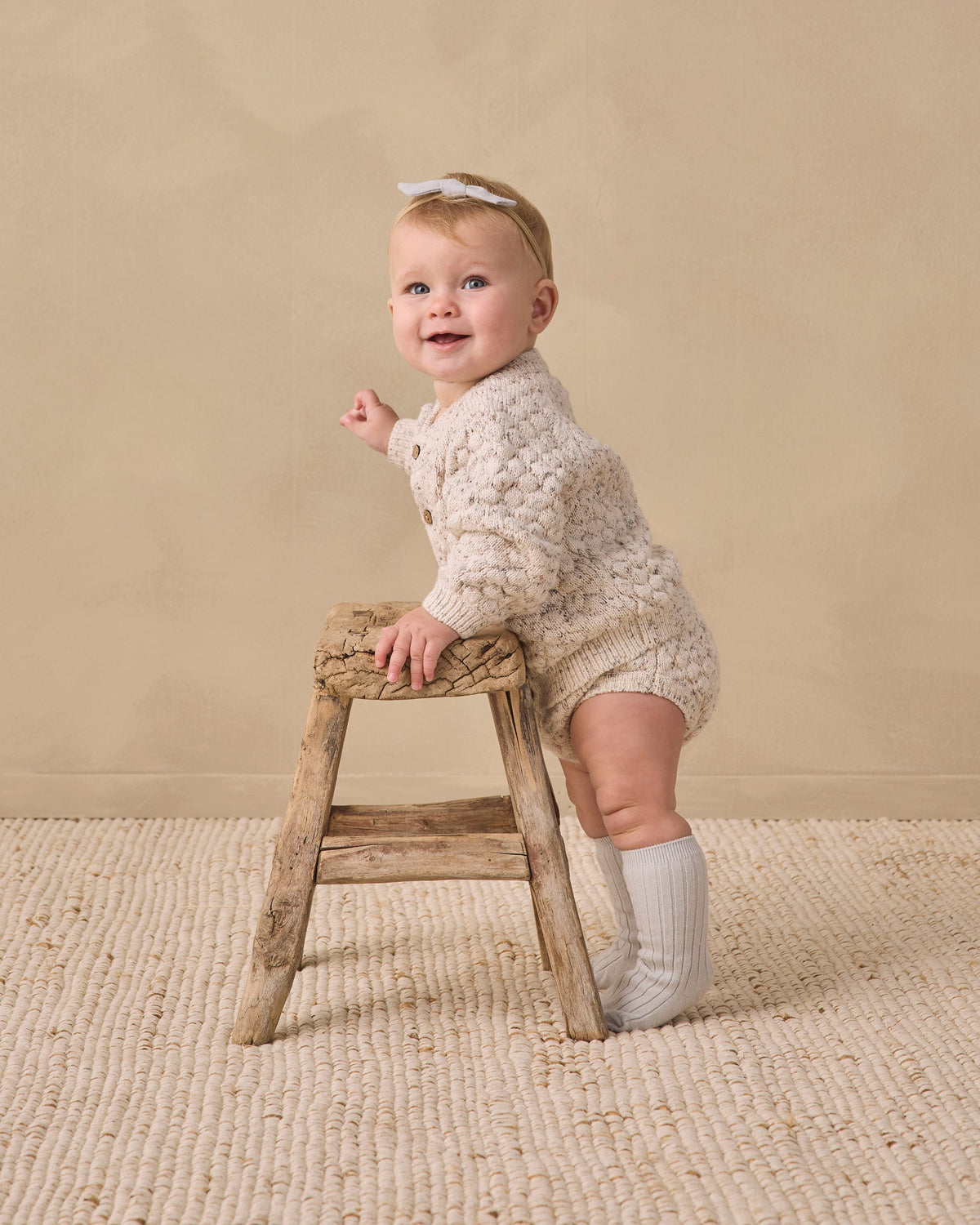 Happy baby in Rylee + Cru Bubble Knit Cardigan holds stool, stands on a textured rug.