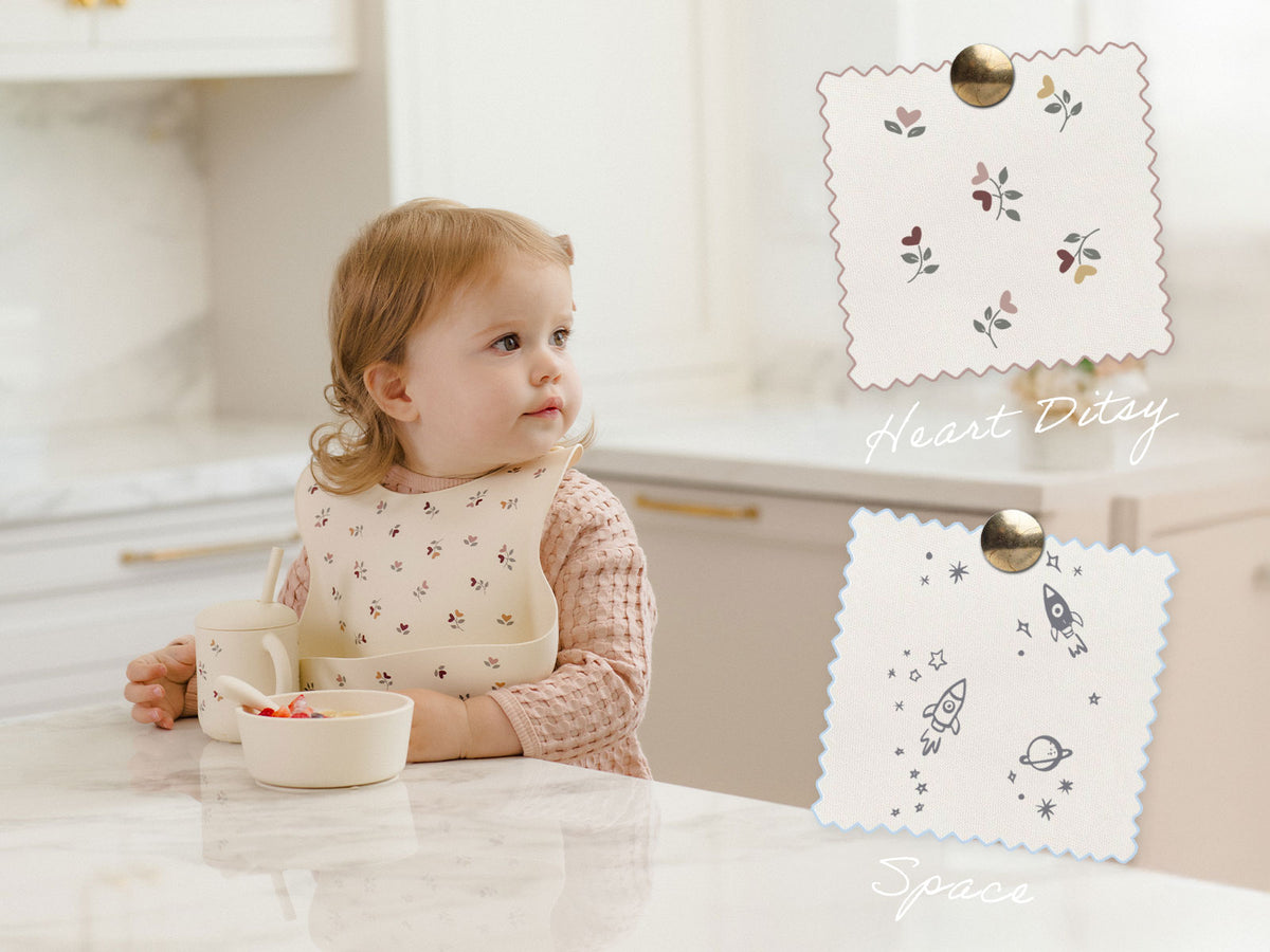 A toddler wearing a cream bib with a small floral print sits at a kitchen counter. Two bib color/style options are shown on the right: one with a scalloped edge, gold clip, and small floral motifs labeled 'Heart Ditty,' and another with a scalloped edge, gold clip, and space-themed illustrations labeled 'Space.