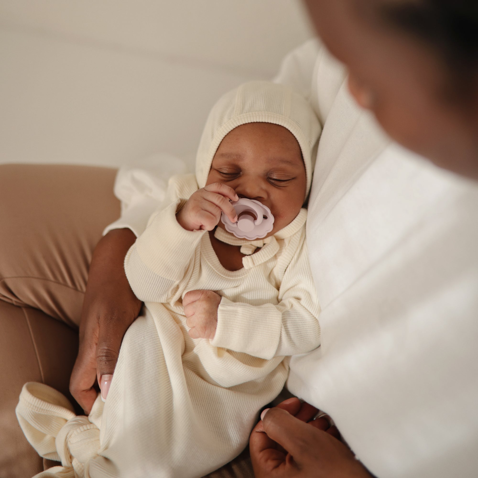 Baby in cream outfit with bonnet, comforted by FRIGG Daisy Natural Rubber Pacifier.