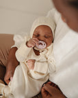 Baby in cream outfit with bonnet, comforted by FRIGG Daisy Natural Rubber Pacifier.