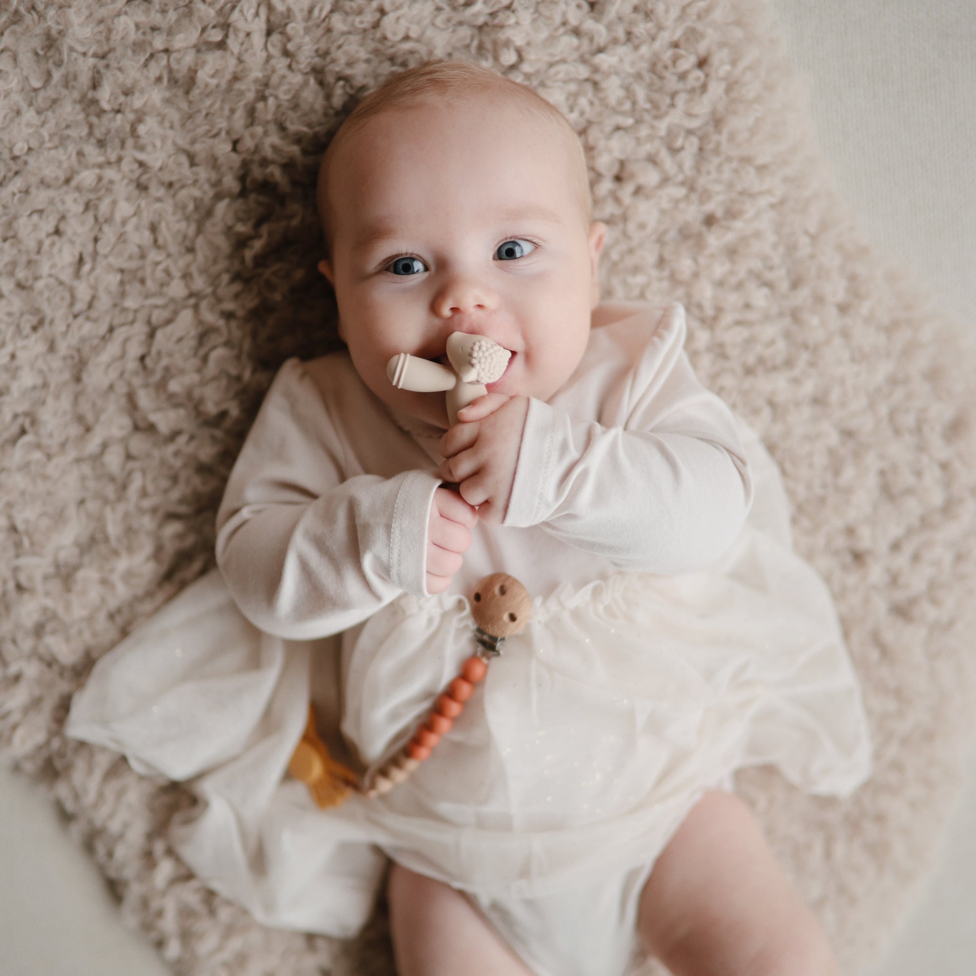 Baby smiles on a soft rug, holding the mushie Figurine Teether.