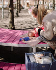 A woman uses the mushie Diaper Caddy while changing her baby on a pink bench outdoors.