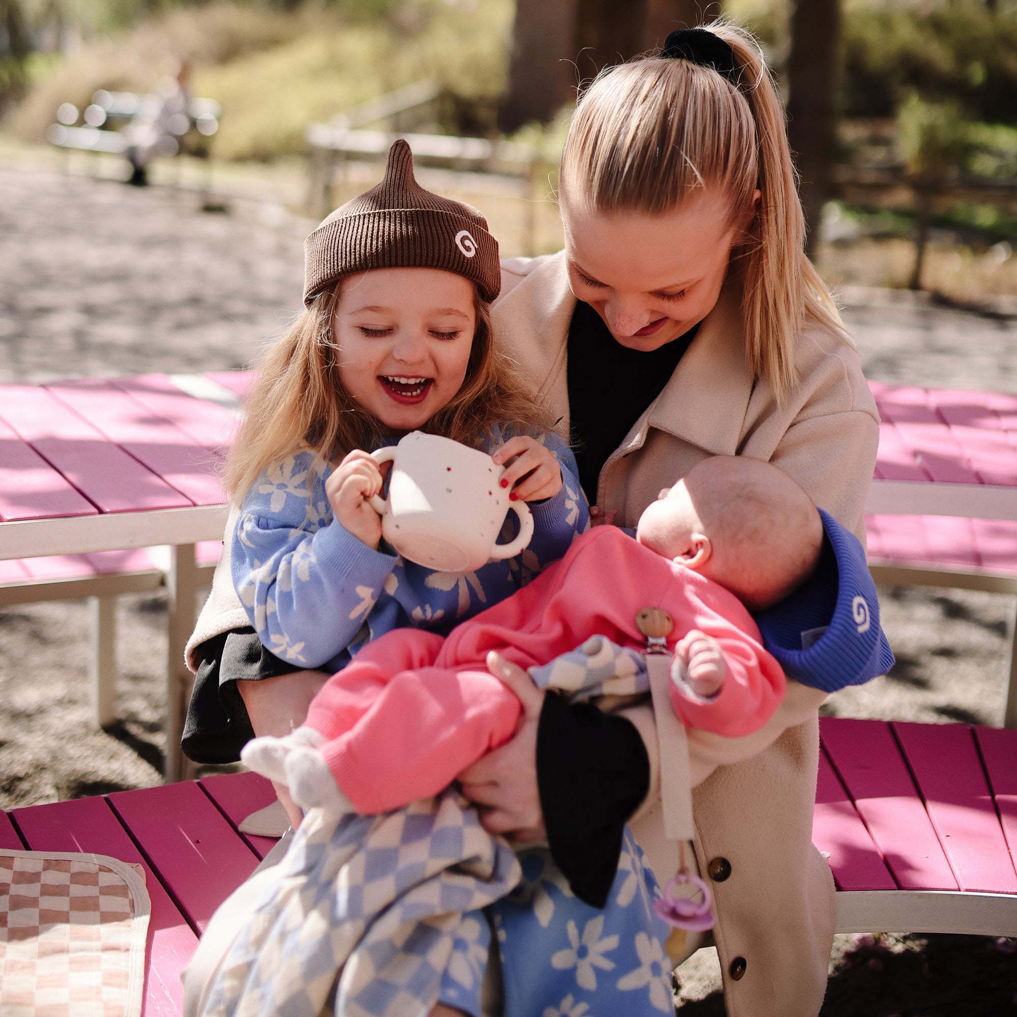 A toddler smiles outside, holding a mushie Snack Cup while a woman holds a baby.