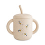 The Mushie Silicone Training Cup + Straw is a beige cup with two handles, a lid, and spout, featuring three bumblebee illustrations and dotted flight paths—ideal as a transition cup for little ones learning to drink on their own.