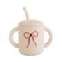 Mushie Silicone Training Cup + Straw in beige with handles, pink bow, and food-grade lid.