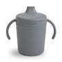 The Mushie Trainer Sippy Cup is gray, non-toxic, and features easy-grip handles with a spout lid. "mushie" is embossed on the front.