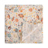 The mushie Organic Cotton Muslin Swaddle Blanket in Space features a colorful space-themed pattern with planets, rockets, stars, and moons on a white background. One corner is folded up to reveal the reverse side.