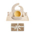 The mushie Stage 1 Bees Feeding Kit—a beige silicone set with bib, bowl, cup, spoon, and fork, all adorned with small floral prints—sits on a matching placemat above a brown gift box tied with white ribbon. Ideal baby mealtime essentials.