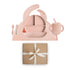 The mushie Stage 2 Blush Feeding Kit features a pink silicone bib, divided plate, bowl, cup with handles, training cup with straw, spoon, and fork on a matching placemat above a brown gift box tied with a white ribbon.
