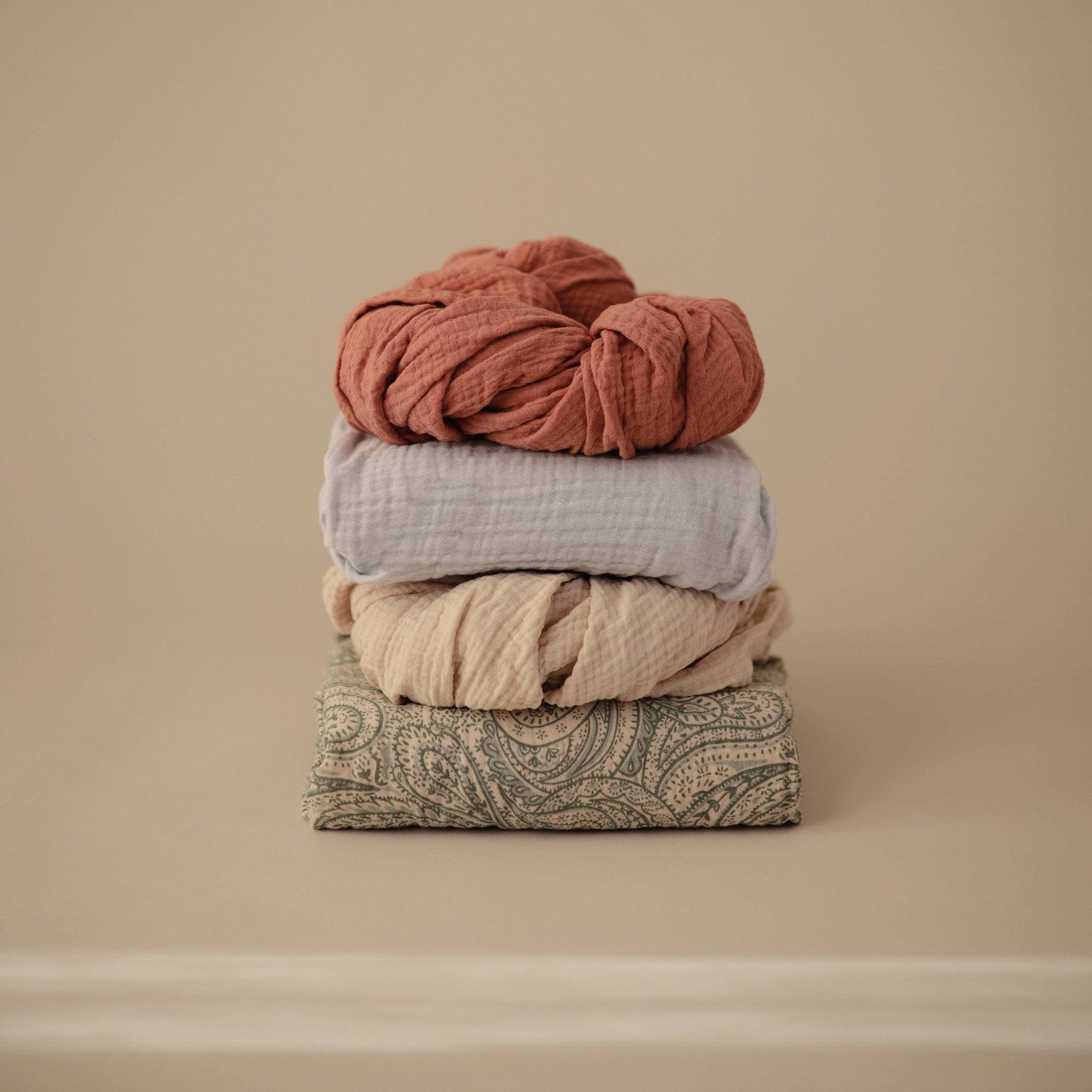 Four mushie Extra Soft Muslin Crib Sheets stacked on a beige background.