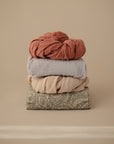 Four mushie Extra Soft Muslin Crib Sheets stacked on a beige background.
