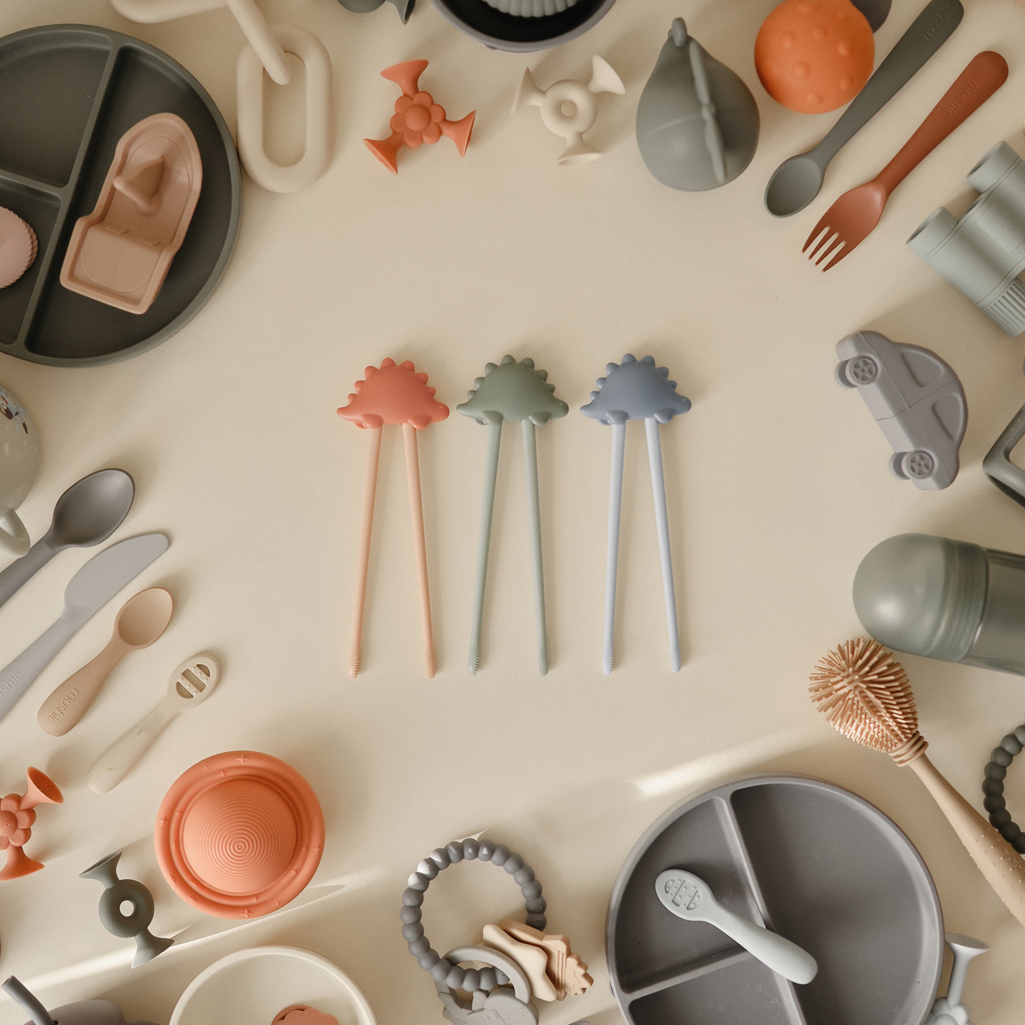 Mushie Training Chopsticks are surrounded by assorted silicone baby utensils and toys.