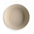 A beige, round, shallow Round Dinnerware Bowl by mushie, made from BPA-free plastic and embossed with the brand name in the center, photographed from above on a white background.