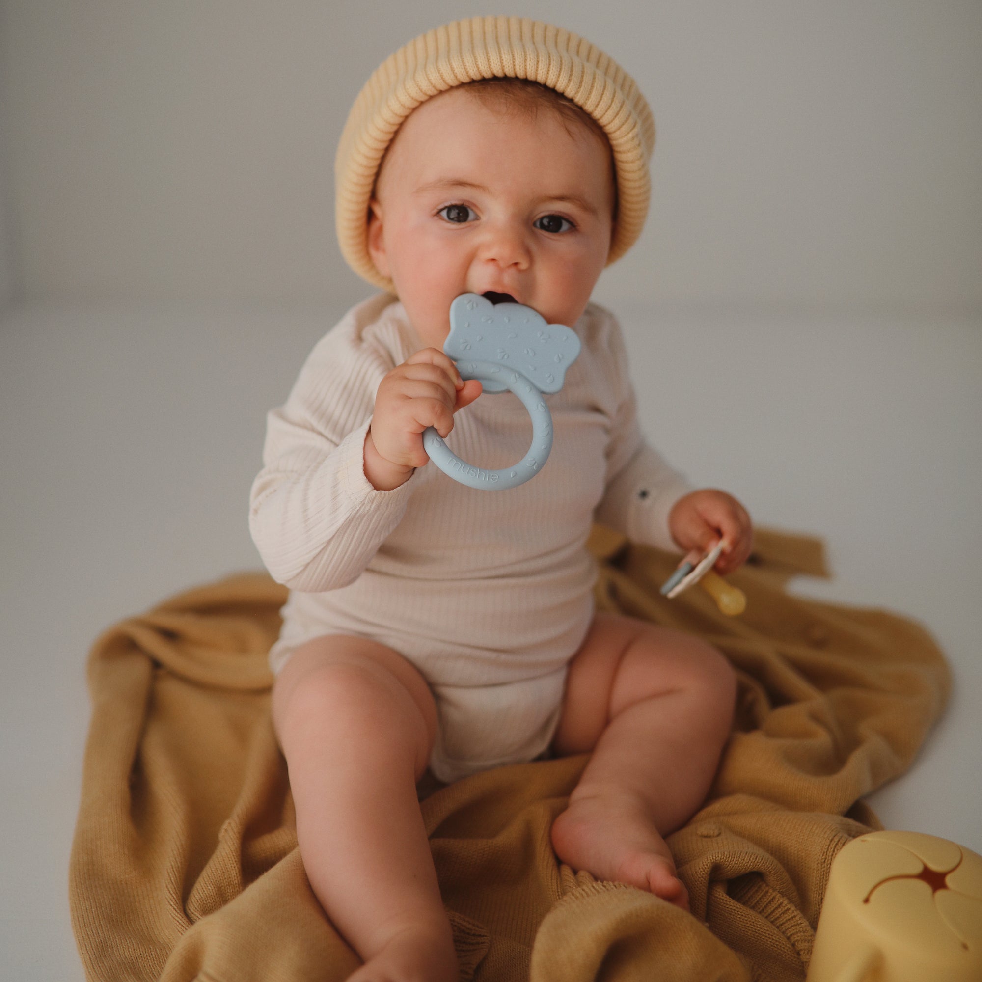 Baby in beige outfit chews on Mushie Animal Ring Teether while sitting on blanket.