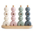 The Mushie Wooden Multi Shape Stacker is an educational toy featuring pastel geometric pieces on four vertical rods set in a light wood base, designed to help develop fine motor skills.