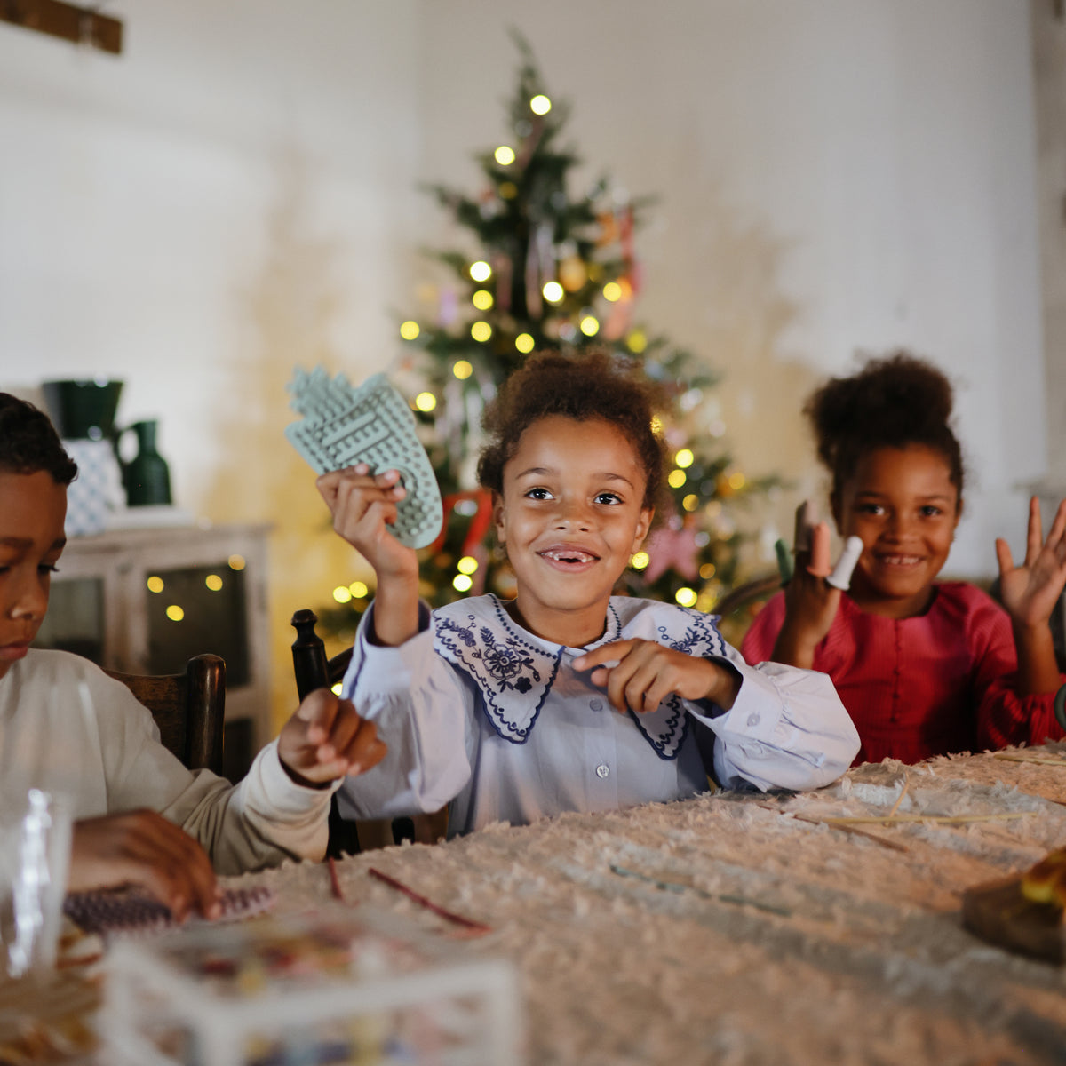 Three kids smile by the tree, each holding a mushie Silicone Sensory Toy at a festive table.