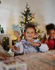 Three kids smile by the tree, each holding a mushie Silicone Sensory Toy at a festive table.