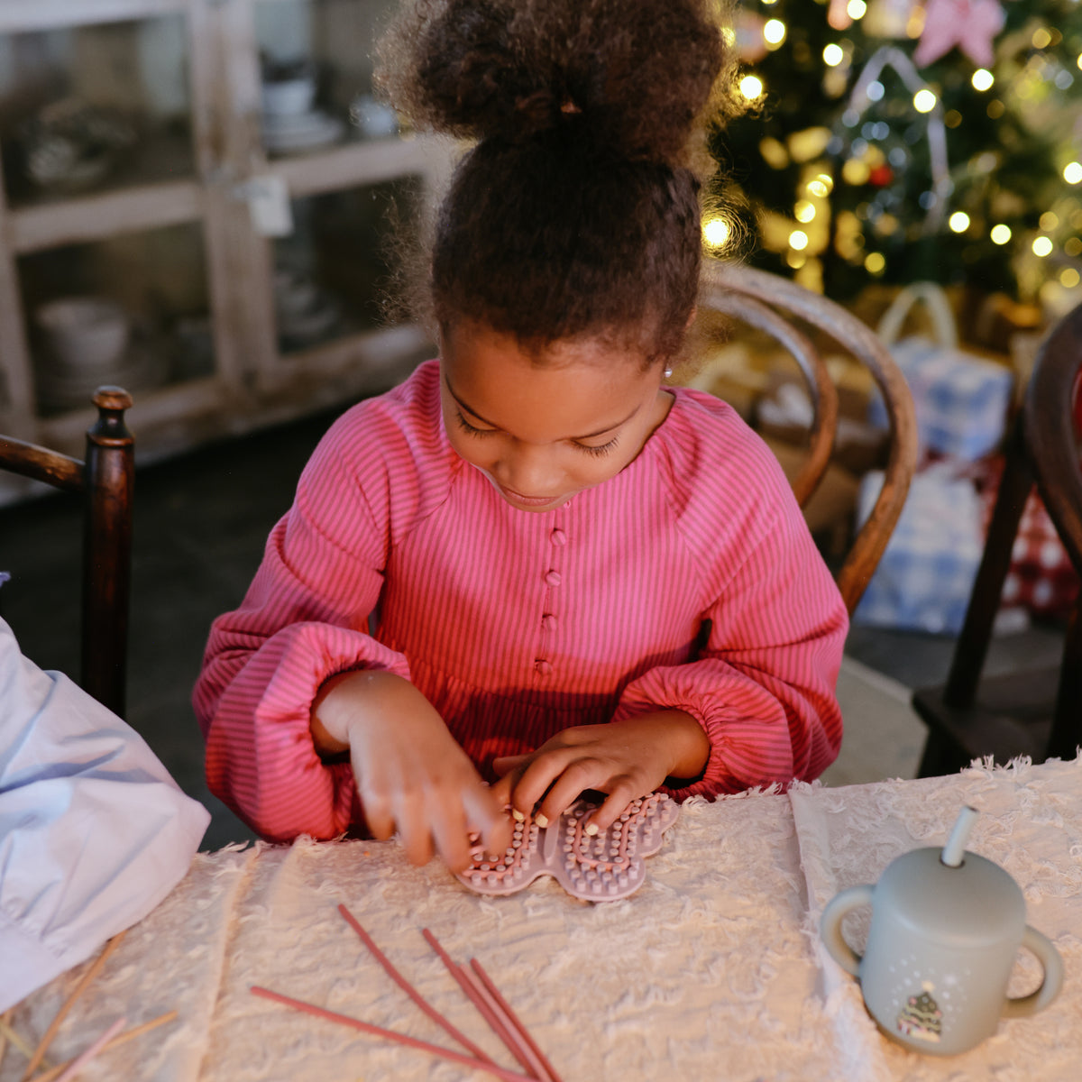 Girl in pink dress plays with mushie Silicone Sensory Toy by the Christmas tree.