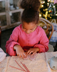 Girl in pink dress plays with mushie Silicone Sensory Toy by the Christmas tree.