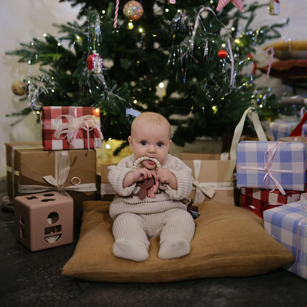 Baby enjoys mushie Playtime Gift Set by the tree, surrounded by gifts and sensory toys.