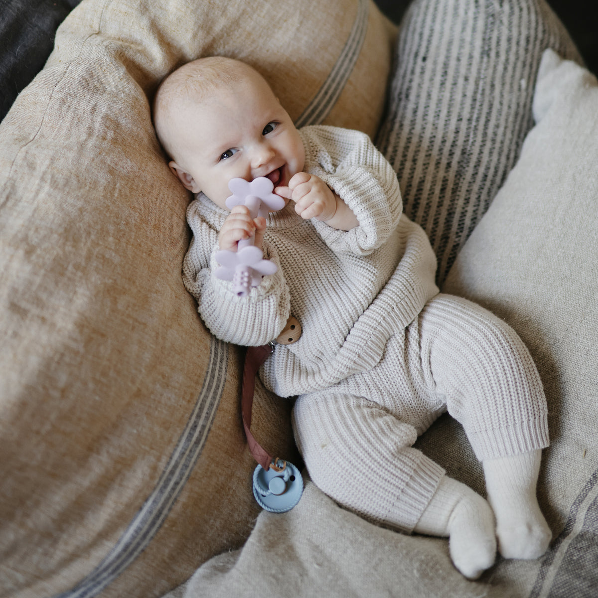 Happy baby in mushie knit outfit with Linen Pacifier Clip 2-Pack, holding flower teether.