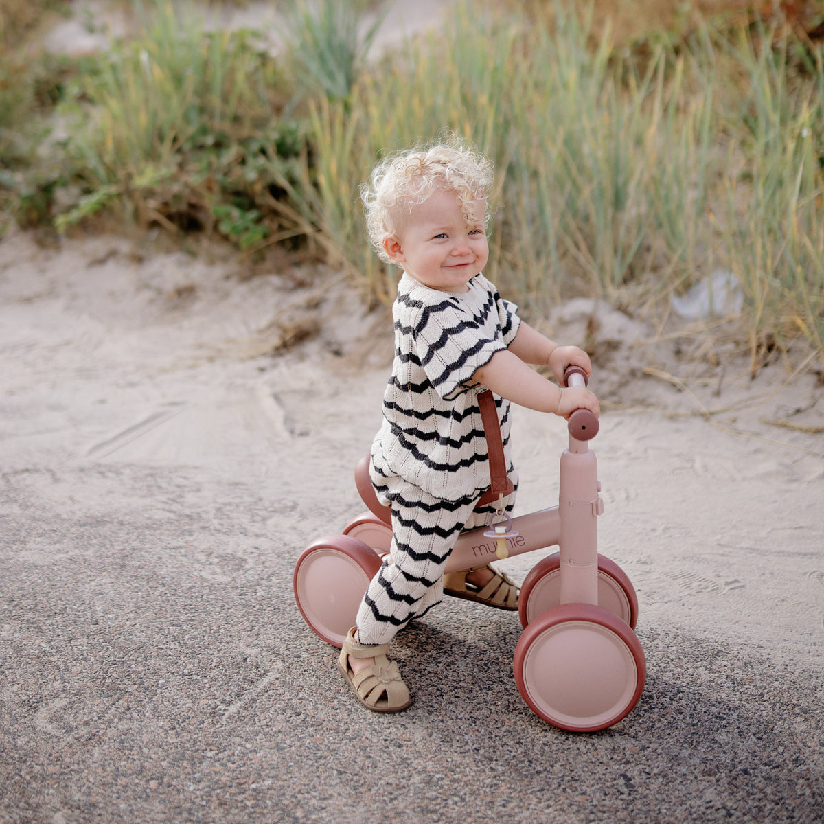 Toddler outdoors in striped outfit with mushie Linen Pacifier Clip 2-Pack, smiling on pink bike.