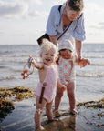 A woman and two toddlers play on the beach, holding mushie Linen Pacifier Clip 2-Pack.