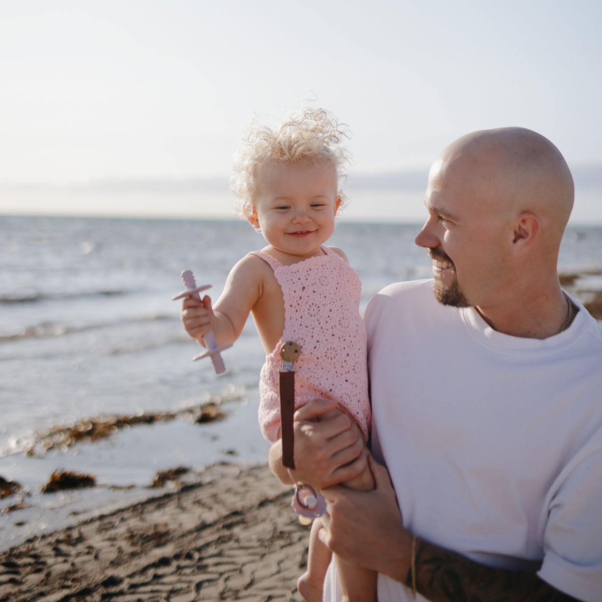 Smiling man holds baby with mushie Linen Pacifier Clip 2-Pack at the beach in sunlight.