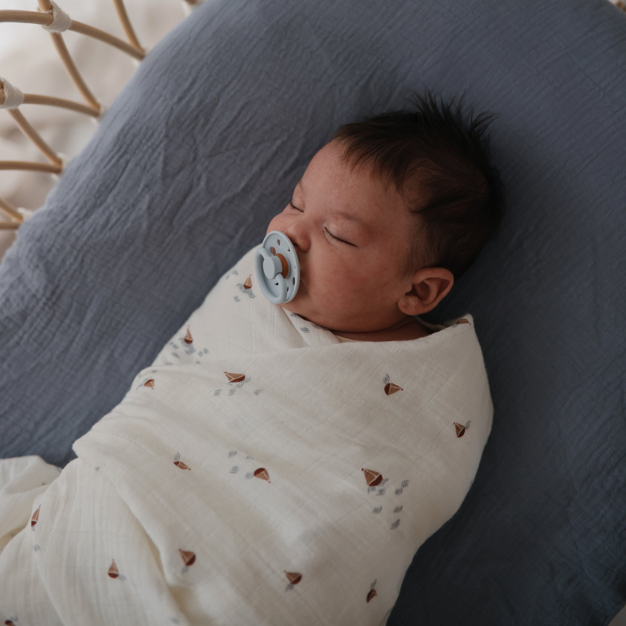 A baby sleeps in a mushie Organic Cotton Muslin Swaddle Blanket with a pacifier on a gray blanket.