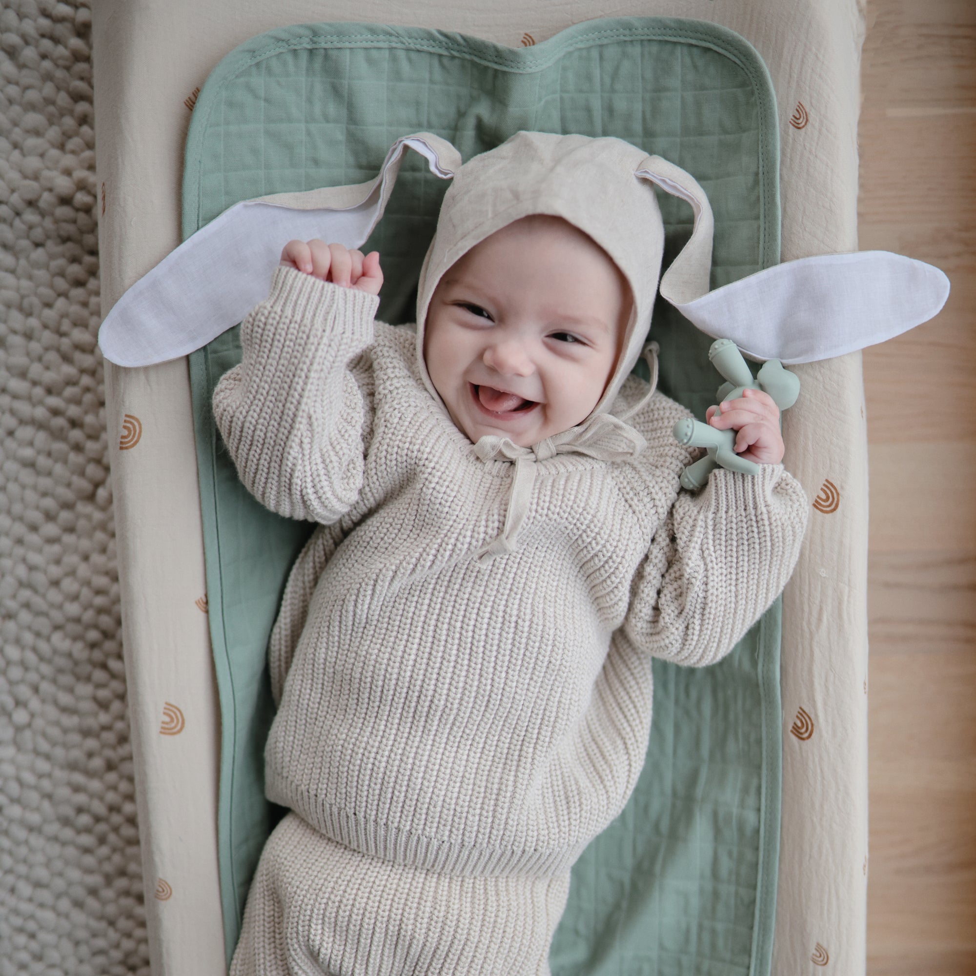 Smiling baby in a bunny-ear hat cuddles the mushie Figurine Teether on a soft blanket.