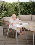 Smiling baby in a high chair with mushie Adjustable Silicone Strap 2-Pack on a patio.