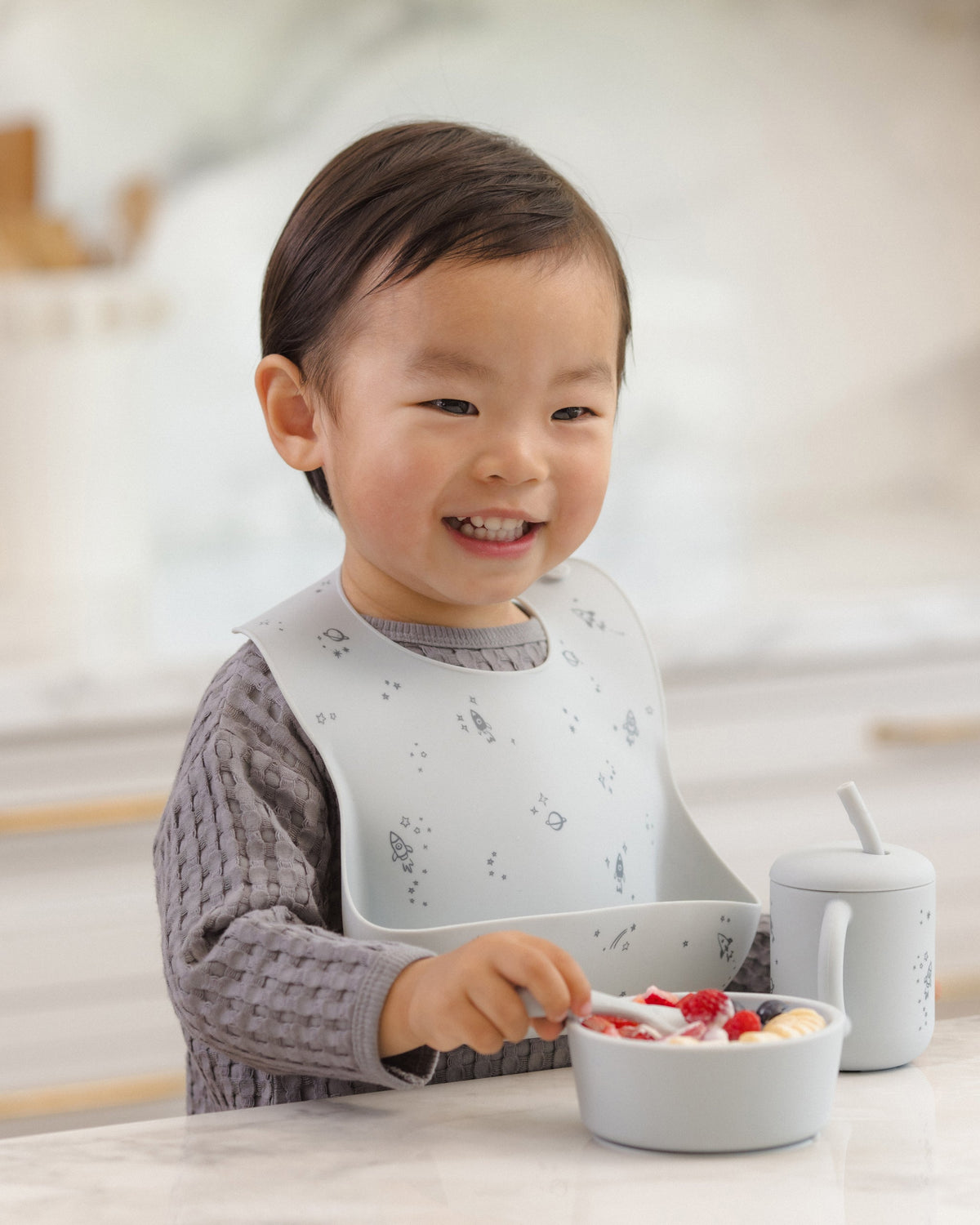 Toddler in Rylee + Cru Feeding Kit Space bib eats fruit, cup nearby on the table.