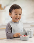 Toddler in Rylee + Cru Feeding Kit Space bib eats fruit, cup nearby on the table.