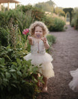 Curly-haired child in a tutu with a FRIGG Daisy Natural Rubber Pacifier and pink flower in the garden.