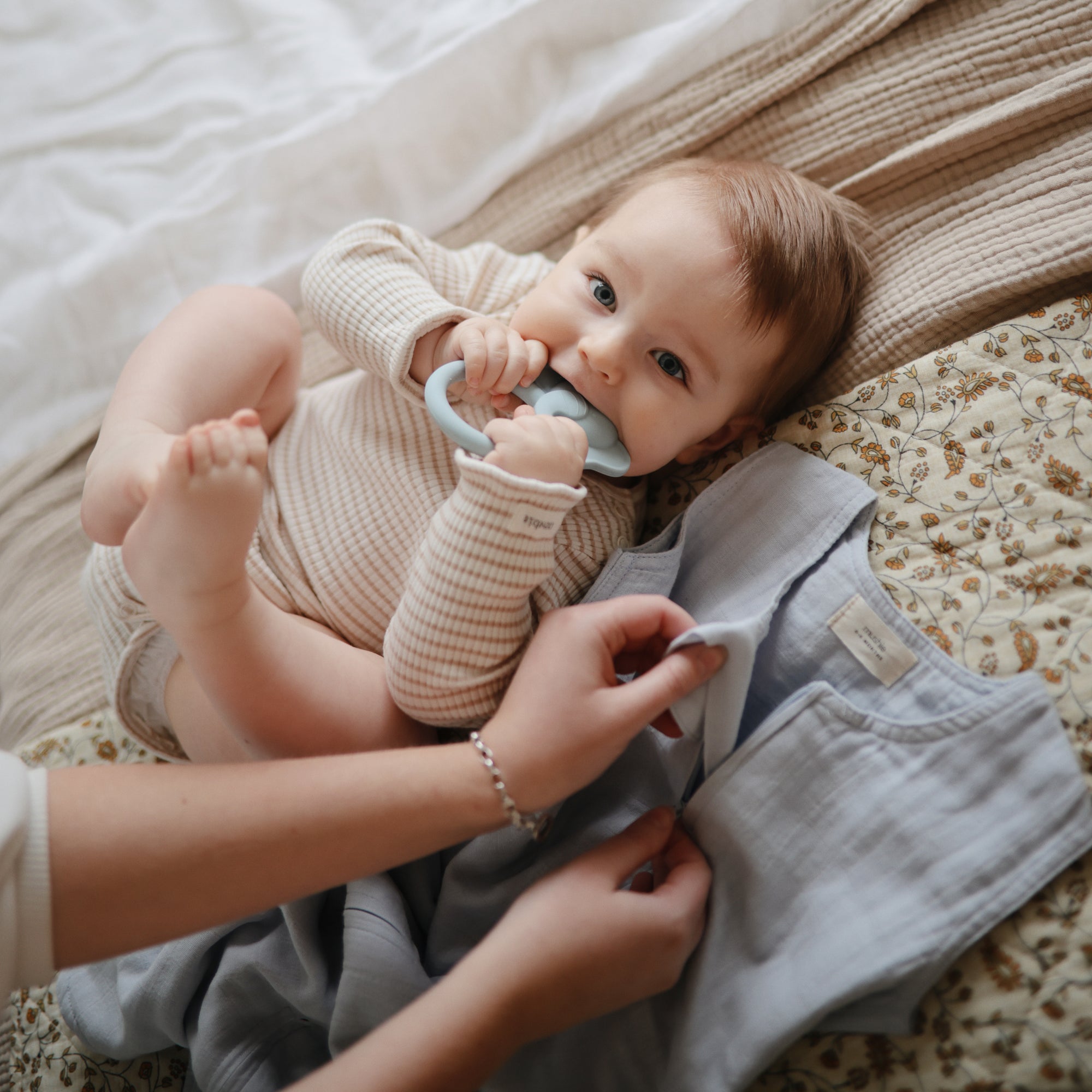 Baby chews Mushie Animal Ring Teether as an adult fastens their blue garment on a blanket.